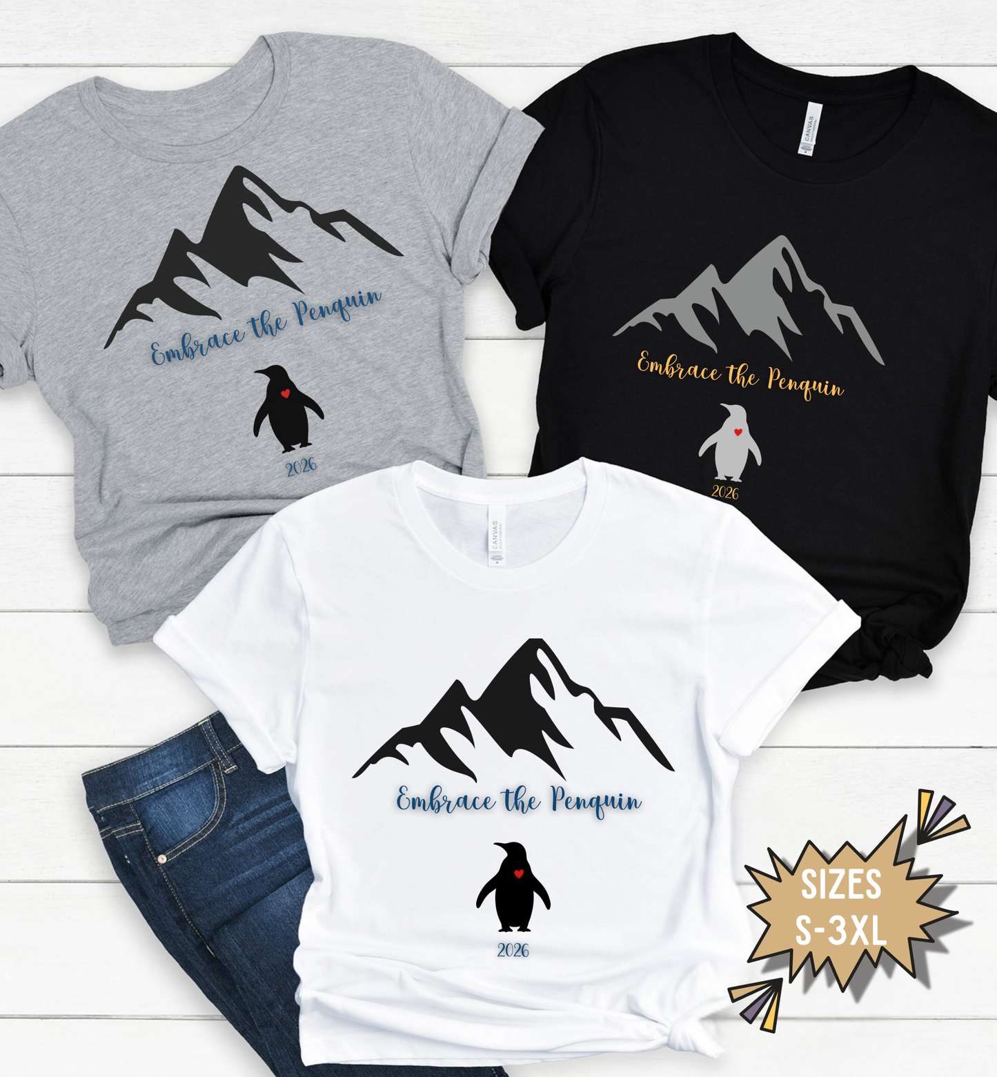Three t-shirts with mountain and penguin designs on a white wooden surface.