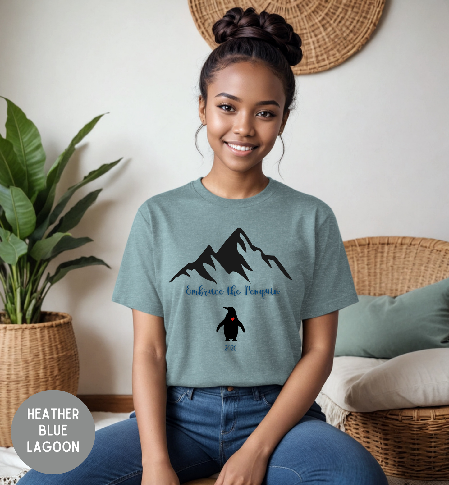 Person wearing a heather blue lagoon t-shirt with a mountain and penguin design, sitting in a cozy room.