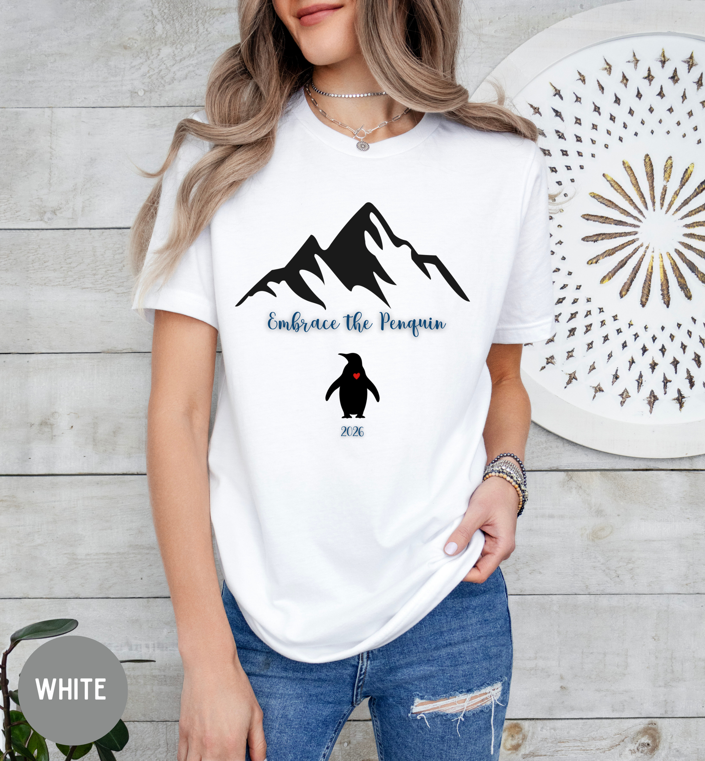 Person wearing a white t-shirt with mountain and penguin design, standing against a wooden wall.