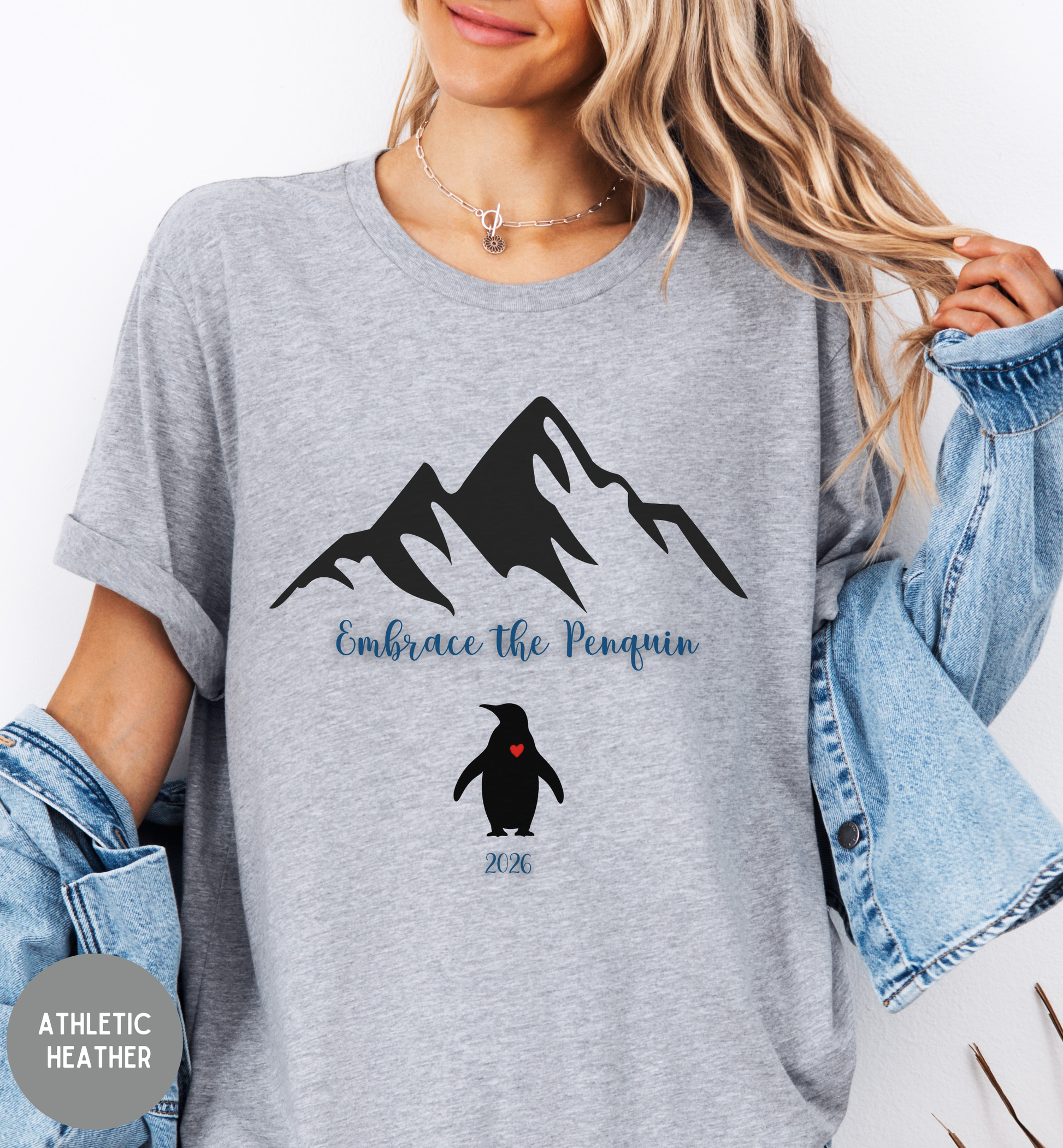Person wearing a gray t-shirt with mountain and penguin design, text 'Embrace the Penguin', and 'Athletic Heather' label.