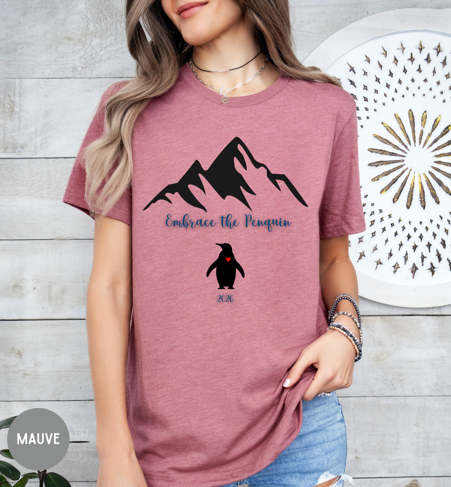 Person wearing a mauve t-shirt with mountain and penguin design, standing against a wooden wall.